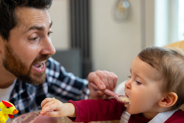 Father feeding his infant baby in home interior teaching toddler kid eating from spoon.