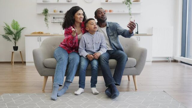 Smiling multicultural family in jeans clothes looking at cell phone camera while seating in lounge on sunny day. Happy husband and wife with preschool son taking video call from grandfather via app.