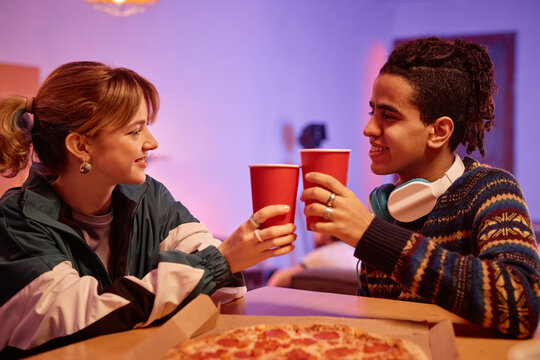 Side view portrait of retro young couple eating pizza and toasting with red paper cups