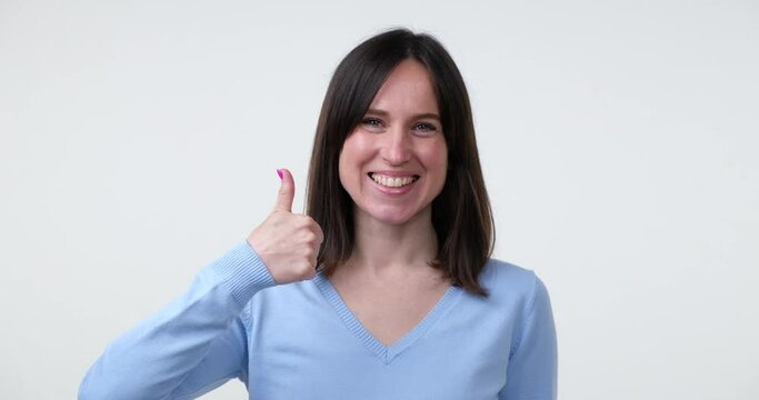Positive Caucasian Woman Showing Thumb Up Gesture. A young woman with a bright smile stands against a white background and gives a thumbs up gesture, indicating approval or liking of something.