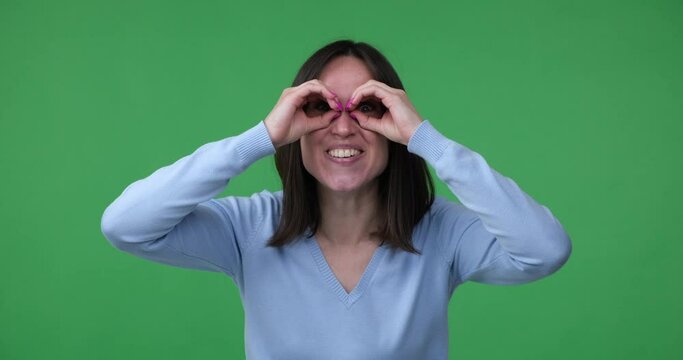 Joyful woman stands on a green screen background, making a binocular gesture with her hands near her eyes. She appears to be searching for something and expresses surprise and amazement