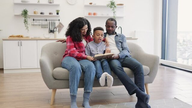 Caring multicultural parents sitting near preschool boy with paper book in hands during summer holidays. Mindful mother checking reading while focused father folowing story characters.