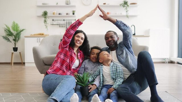 Joyful multicultural couple holding hands high in air while happy boys with plant pot sitting on floor im front of sofa. Creative adults building alive toy house with smiling children inside.