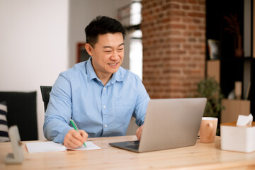 Happy asian middle aged man freelancer using laptop and taking notes while working at home, sitting at desk, copy space
