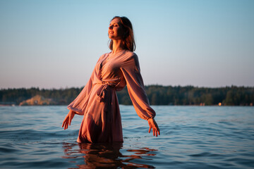 Beautiful caucasian woman in pink dress in water on the sunset