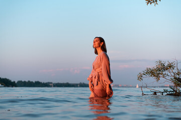 Beautiful caucasian woman in pink dress in water on the sunset