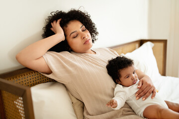 Sad tired bored black millennial mom holds in her arms small baby, sleeping on bed in bedroom