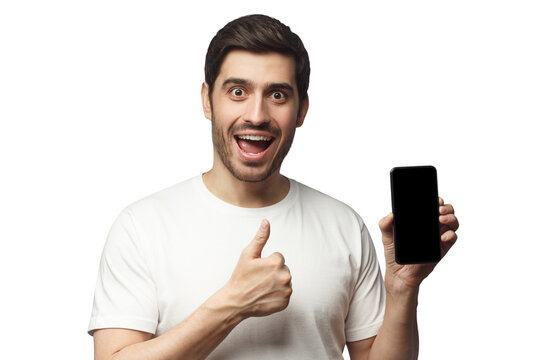 Excited screaming WOW young man in white t-shirt presenting blank black smartphone screen and showing thumbs up gesture