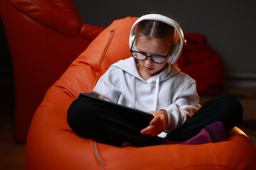 Concentrated teen girl in headphones plays game, communicate on internet social media, listens to music or online studies at home on digital tablet sitting on sofa. Children tech addiction concept