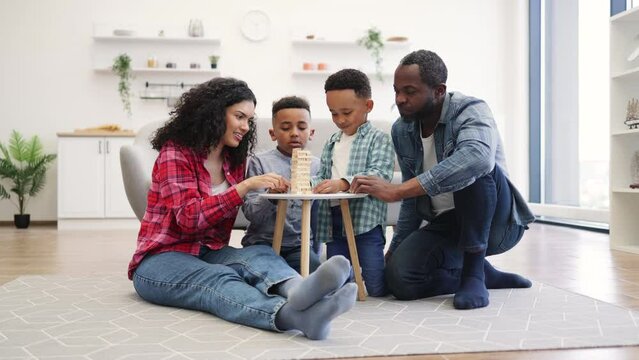 Friendly multiethnic family gathering together around coffee table on room floor at noon. Happy kids and involved parents building high tower of wooden blocks on floor of kitchen.