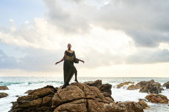 Trans sexual black person poses on scenic ocean beach like a goddess. Lgbtq ethnic fashion model in long posh dress and accessories stands on rocks by sea against moody sky in dull cloudy weather