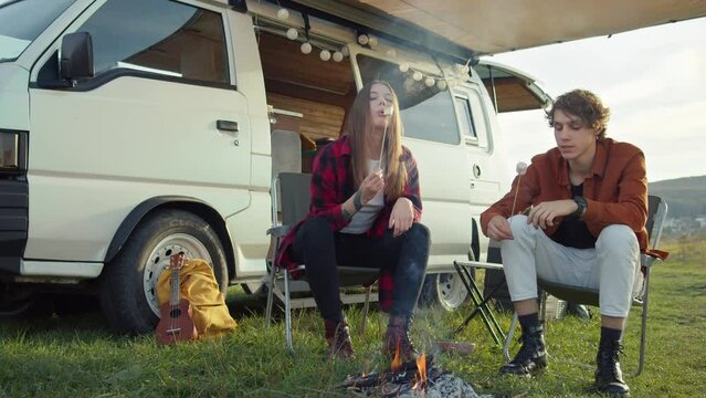 Long-haired girl blows on a hot marshmallow on a stick near a campfire. Young caucasian couple roasting marshmallows at camping. The romantic road trip, outdoor weekend, picnic in nature