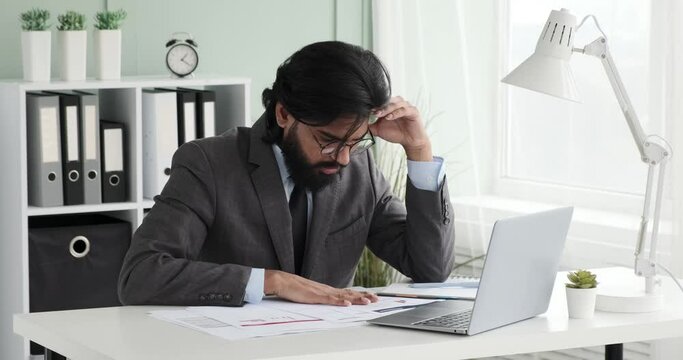 Indian businessman wearing a suit is seen checking papers on his laptop. He then removes his glasses and massages his temples, showing signs of a headache.