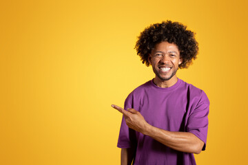 Cheerful mature black curly guy in violet t-shirt pointing finger at free space, isolated on yellow background