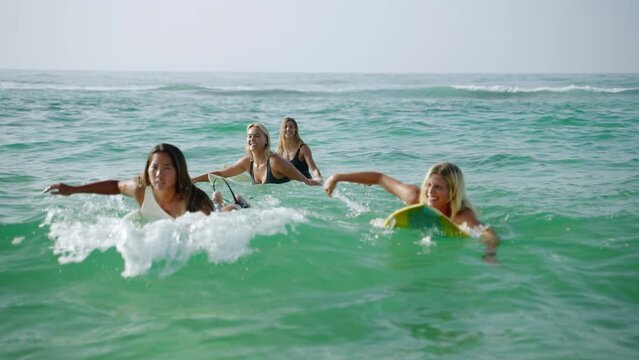Young multiracial women surfers rowing on surfboards and riding waves in the sea at tropical beach at sunrise. Girl friends enjoy outdoor lifestyle and water sport surfing on summer vacation at dawn