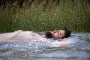 young bearded man lying in the river blissfully with vegetation in the back