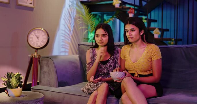 Indian asian two young female sitting on sofa hold bowl watching TV sad movie eat popcorn snacks late night at home. Upset teen women girl wipe tears look dramatic film cinema spend weekend time