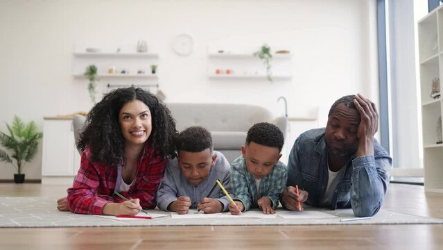 Four african american people using colored pencils while lying on warm wooden floor in kitchen interior. Focused mother and father helping little sons with drawing during summer vocations at home.