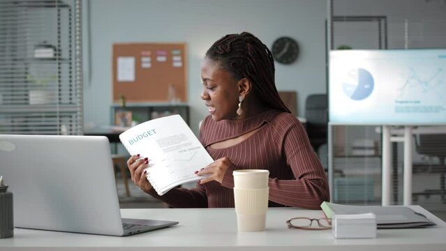Waist up of young Black woman with financial report in hands making conference business call in bright office