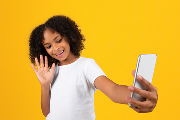 Happy adolescent curly black girl in white t-shirt waving hand in smartphone, has video call