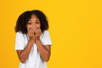 Glad shocked pretty teen curly girl in white t-shirt bites her fingers, isolated yellow studio background