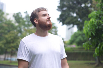 Positive young man smiling during the rain in the park. Cheerful male enjoying the rain outdoors. Guy looking up and catching the rain drop with hands. Breath deep and relax