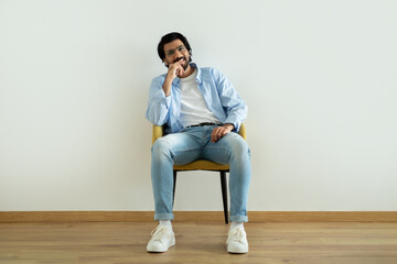 Smiling pensive millennial middle eastern guy in glasses sits on armchair with laptop in room