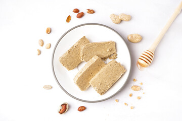 Plate with slices of tasty halva, peanuts and honey dipper on white background