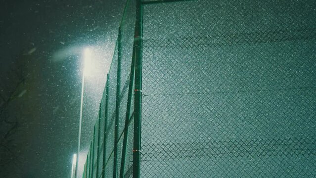 View of green metal mesh fence in night setting and heavy snowfall. Gloming artificial illumination. bright white lite from the lamp. Late working sports field in winter season
