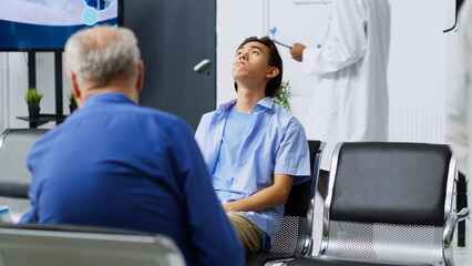 Fototapeta na wymiar Tired asian patient being nervous during checkup visit appointment while waiting for specialist to discuss medication treatment. Young adult sitting on chair in hospital waiting area.