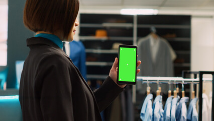 Young assistant using blank greenscreen template, showing chroma key display in trendy clothing store. Asian worker using isolated mockup copyspace on modern smartphone, shopping center.