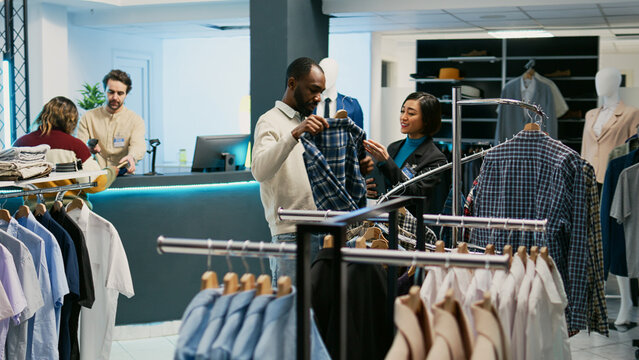 Asian woman showing shirts to male client in mall store, searching for discount merchandise in shopping center. Female shop assistant looking at trendy clothes on display in boutique shop.