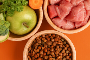Bowls with dry pet food, raw meat and natural products on orange background, closeup
