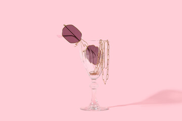 Glass with stylish sunglasses and chain necklace on pink background