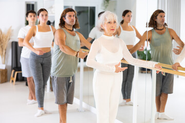 Slim aged woman practicing choreography at ballet barre in gym room during training session
