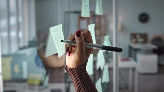 Close up of unrecognizable businesswoman writing on sticker note attached to glass window while planning work in office