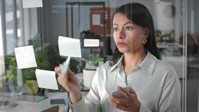 Chest up of young confident Biracial businesswoman writing notes on sticker notes and putting them on glass window while planning work in office