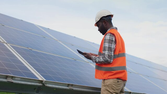 Portrait of African American engineer working at production of alternative energy sources. Industrial employee in helmet and protective vest holding digital tablet for counting and making tasks.