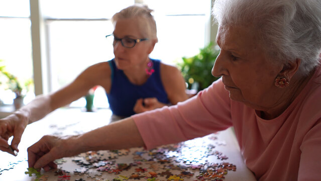 Closeup of happy pretty elderly woman organizing puzzle pieces while daughter moves pieces to fit together. Concept of spending time with loved ones.