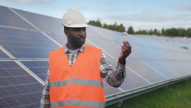 Concept of green electricity. Portrait of African-American employee working in production of solar panels. Male dressed in hard hat is holding light bulb and looking into camera. Alternative sources