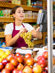 Smiling european female seller weighing fresh grape in supermarket