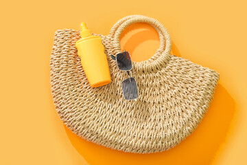 Bottle of sunscreen cream with sunglasses and wicker bag on orange background