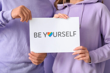 Young couple holding paper with text BE YOURSELF on lilac background, closeup. Pride Month