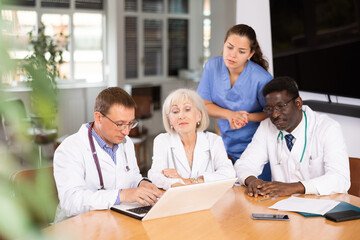 Three interested doctors sitting and a nurse standing at table searching information by means of notebook
