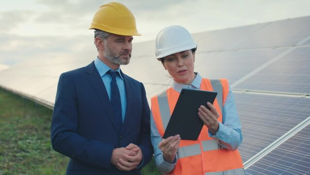 Man and woman in hard hats discuss work tasks on solar panel background outside. Female ecological engineer holds digital tablet and talking with investor. Concept of green electricity.