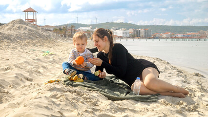 Fototapeta premium Cute baby boy with mother relaxing on the sandy beach and having a picnic. Family vacation and weekend on nature.