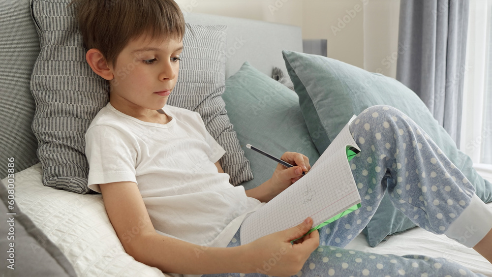Wall mural portrai of little boyworking on his homework in bed, writing in a notebook with concentration. impor