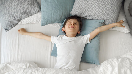 Cute little boy lying in bed, stretching hands and yawn. Child waking up in bed at morning.