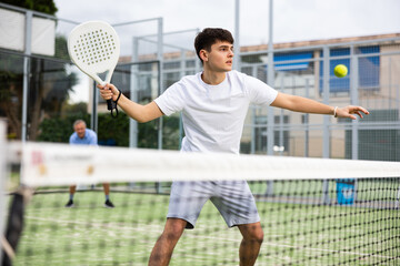 Portrait of emotional determined young guy playing padel tennis on open court in summer, swinging racket to return ball over net. Sportsman ready to hit volley - obrazy, fototapety, plakaty