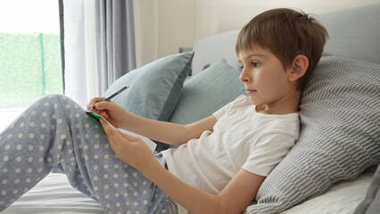 Little boy lying in bed, writing in notebook and ding homework. Concept of education, child...